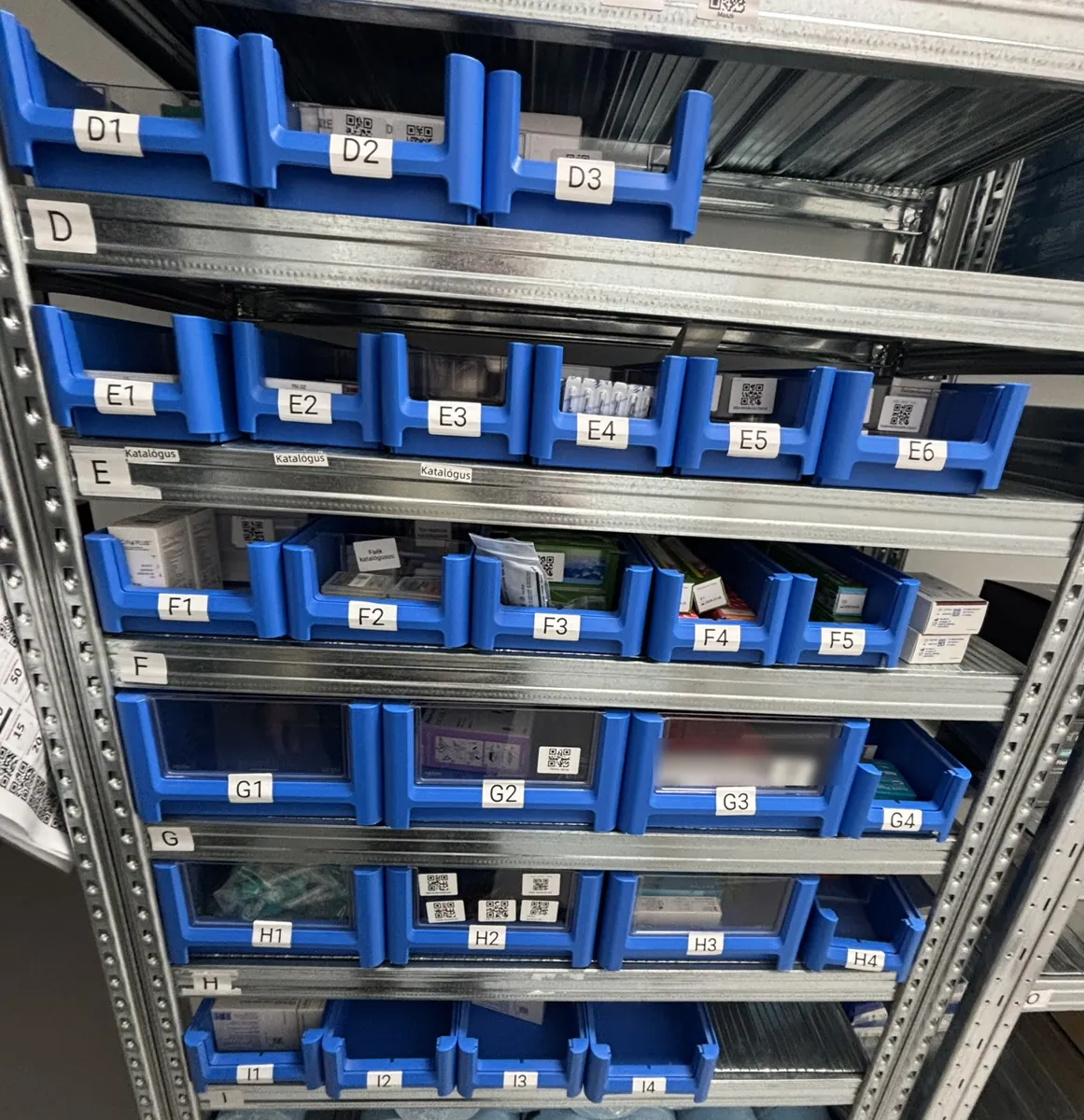 Fém raktárpolc kék dobozokkal, minden dobozon polckód (D1-től I4-ig), a tételeknél QR-címkék